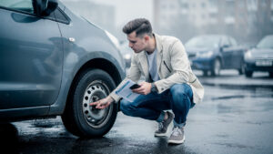 Comment trouver une voiture d’occasion fiable avec un petit budget Un jeune homme inspecte une petite voiture d’occasion sur un parking, tenant un carnet d’entretien et un smartphone.